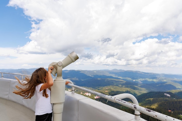 Chica mirando paisajes de las montañas Ródope y el cielo nublado a través del telescopio en la torre de observación en la parte superior de Snezhana