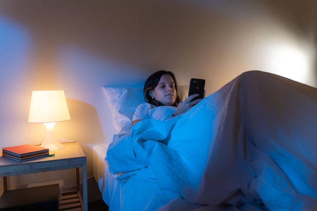 Chica mirando la perdición de su teléfono inteligente desplazándose en la cama en medio de la tecnología nocturna en la cama