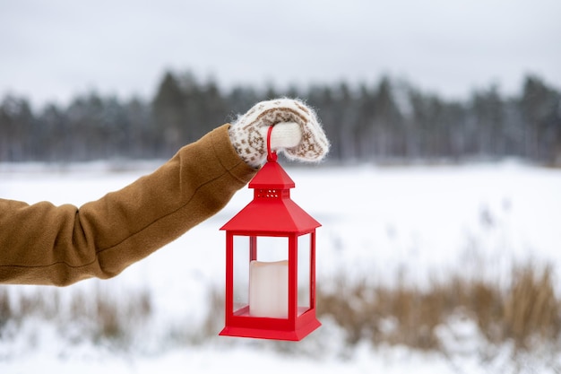 Una chica con mitones de lana sostiene una linterna roja en sus manos con una vela en el bosque de invierno