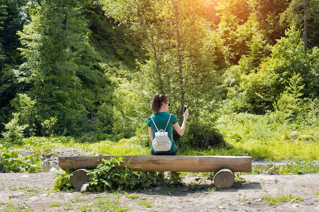Chica con una mochila cerca del bosque toma una foto en un teléfono inteligente. Vista trasera. Día soleado de verano