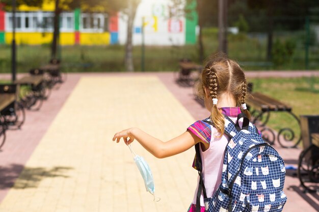 Chica con una mochila cerca de la escuela después de clases con la máscara médica quitada