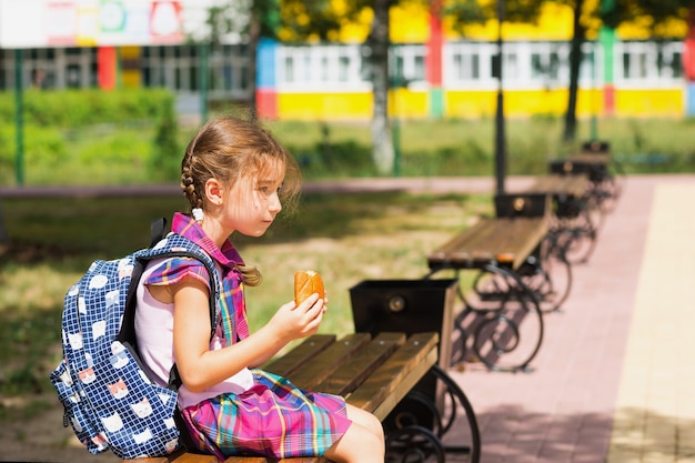 Chica con mochila sentada en un banco y comiendo un pastel cerca de la escuela. Un refrigerio rápido con un panecillo, comida poco saludable, almuerzo de casa. De vuelta a la escuela. Educación, clases de primaria, 1 de septiembre