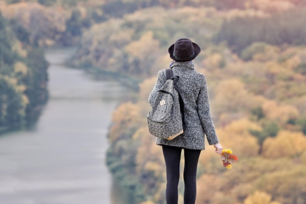 Chica con una mochila y un sombrero de pie en una colina Río y bosque debajo Vista posterior