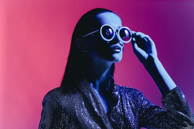 Chica de moda con cabello largo y gafas de sol redondas con un vestido negro brillante posa en luz de neón en el estudio.