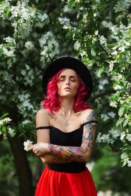 Chica de moda con cabello teñido de rojo brillante en flores de manzana y lilas. Color creativo rosa brillante, colorista. Mujer camina en un parque disfruta de la primavera