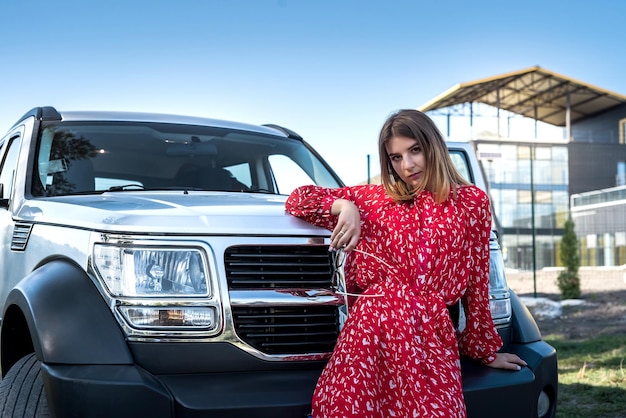 Chica de moda con elegante vestido rojo parada cerca de su auto moderno y relajándose en la naturaleza en el día de verano