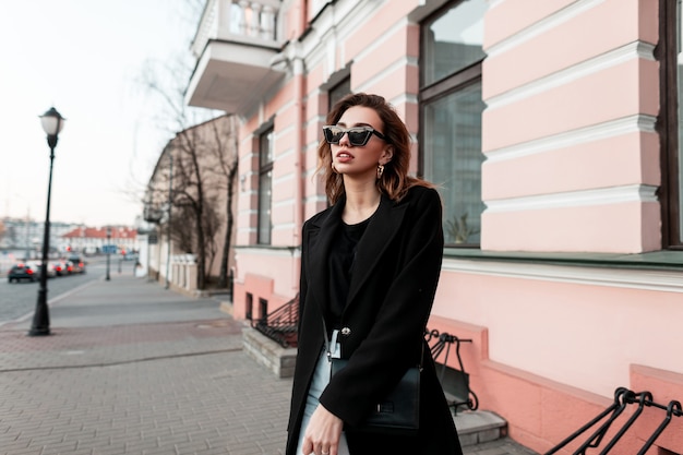 Chica de moda con gafas de sol de moda en ropa negra elegante camina al aire libre