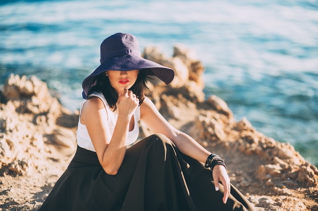 chica de moda en un gran sombrero posando sobre una roca en la playa