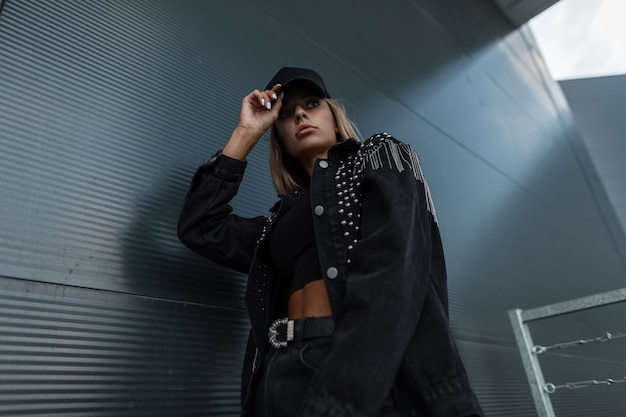 Chica de moda modelo fresco con una gorra negra con una chaqueta de mezclilla negra de moda con una camiseta y jeans posando cerca de una pared de metal oscuro en la ciudad