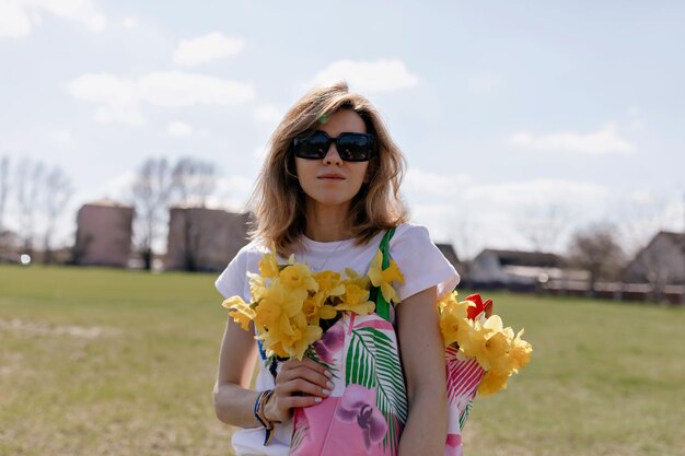 Chica de moda con peinado corto con gafas de sol sostiene una bolsa brillante con flores y posa sobre un fondo natural en un día cálido y soleado