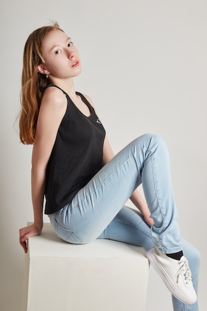 Chica de moda con el pelo largo en ropa casual sentado en un cubo blanco y posando. Hermosa niña pequeña.