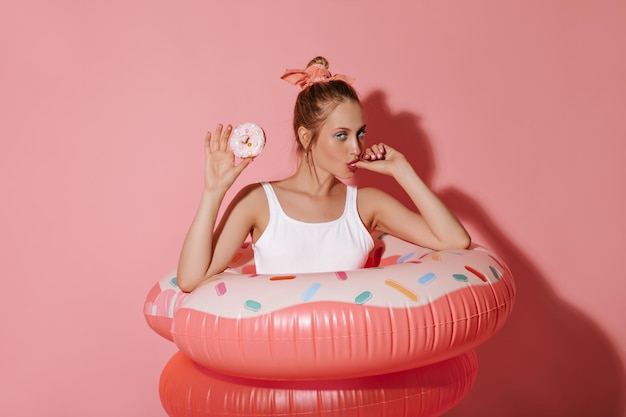 Chica de moda en traje de baño moderno blanco posando con donut y grandes anillos de natación y mirando a la cámara sobre fondo rosa