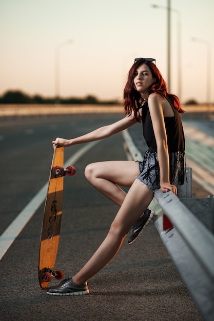 Chica de moda urbana con longboard posando al aire libre en la carretera al atardecer.