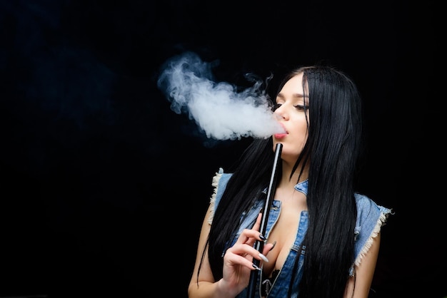 Chica de moda vapeando Nube blanca de humo Vapear es sexy Adicción a la nicotina Morena glamorosa fumando dispositivo de vapeo fondo negro Chica vapeando Descansa y relájate Barra de narguile Cigarrillo electrónico