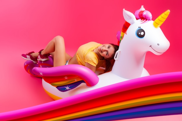 Foto chica de moda de verano, humor rosado y alegre. hermosa mujer delgada en ropa de verano, divirtiéndose y disfrutando de la vida en un unicornio inflable blanco