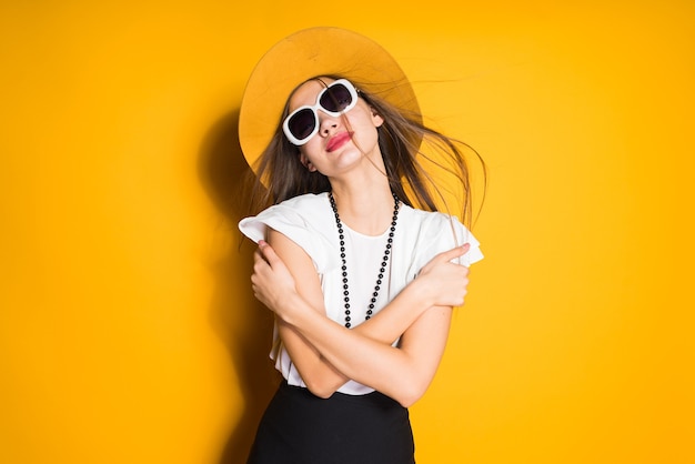 Chica modelo atractiva confiada en sombrero de moda y gafas de sol posando sobre fondo amarillo