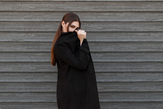 Chica modelo elegante con un abrigo de moda negro cubre su rostro cerca de una pared de madera