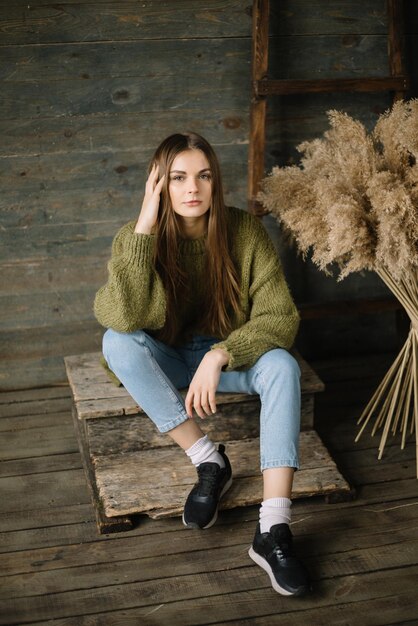 Chica modelo de ensueño de cabello castaño de moda sentada en un estudio rústico de piso de fondo de madera en suéter de punto verde oscuro y jeans azules Brazo de pose de moda cerca de la cabeza mirando a la cámara