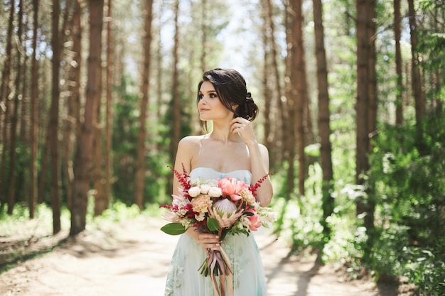 Chica modelo con maquillaje brillante en un vestido con el ramo de flores en la mano posando en el bosque en el día soleado