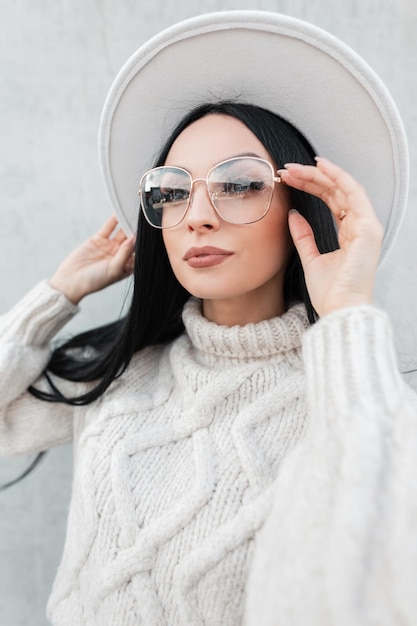 Chica modelo de moda con un estilo hermoso en un moderno suéter de punto blanco y sombrero que lleva gafas de moda vintage contra un fondo gris en la ciudad