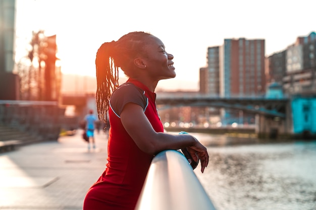 Foto chica modelo con piel negra y cabello estilo afro posando sonriendo aferrándose a la barandilla de un puente junto al río de una ciudad en una hermosa puesta de sol con colores cálidos en un vestido rojo