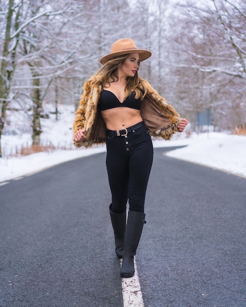 Chica modelo rubia disfrutando en lencería, un sombrero de vaquero y un suéter de leopardo en medio de la carretera con árboles congelados en el fondo, estilo de vida invernal