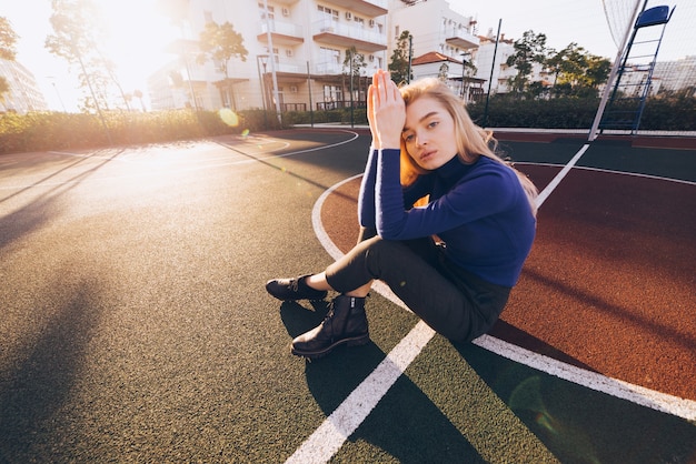 Chica modelo rubia joven elegante en suéter azul posando en el campo de deportes en el sol de la mañana