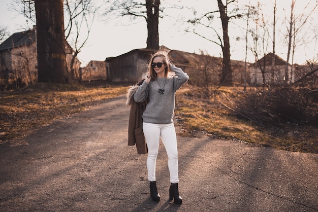 Chica modelo rubia en pantalón blanco, gafas de sol, abrigo marrón