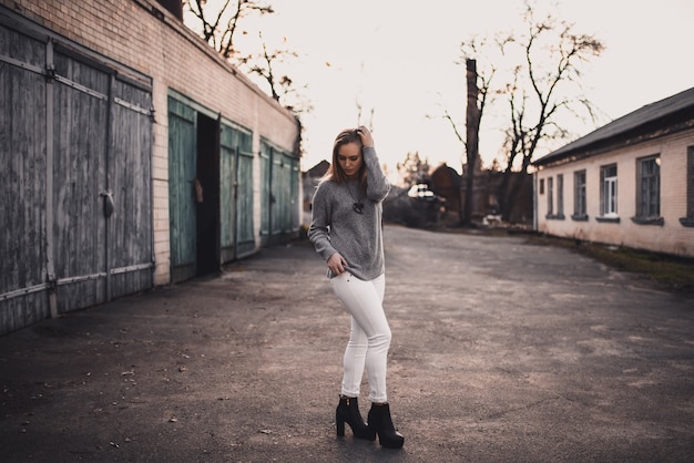 Chica modelo rubia con pantalón blanco, suéter gris y botas negras