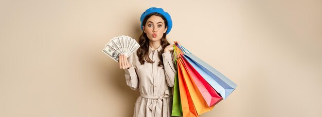 Chica moderna con estilo que siente satisfacción mientras compra posando con dólares de dinero y bolsas de compras beig