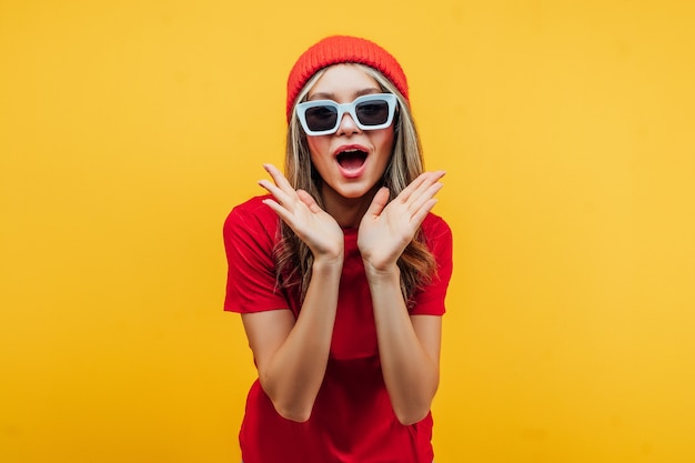 Chica moderna con gafas y un sombrero rojo sobre fondo amarillo, hace una cara de sorpresa, con gafas de sol.