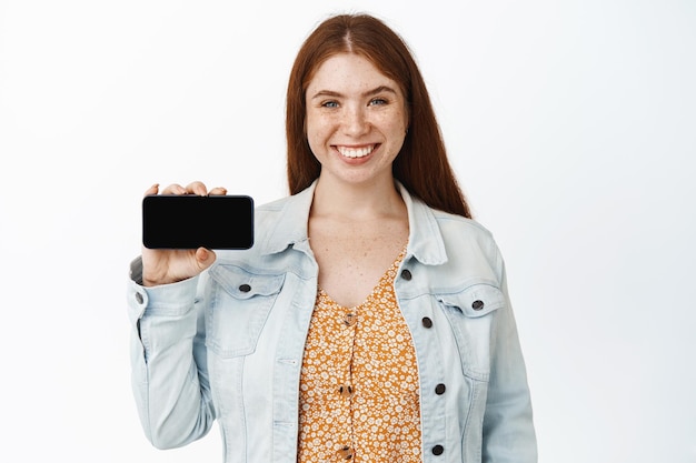 Chica moderna sonriente con cabello rojo sosteniendo el teléfono horizontalmente mostrando la pantalla y la interfaz de la aplicación sonriente en el fondo blanco del teléfono inteligente