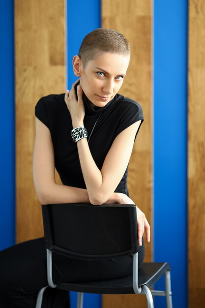 Chica moderna en un vestido negro se sienta en una silla en la pared con tablas de madera.