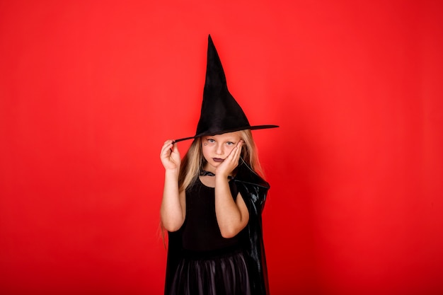 Chica molesta en un disfraz de bruja con un sombrero en una pared roja con una copia del espacio