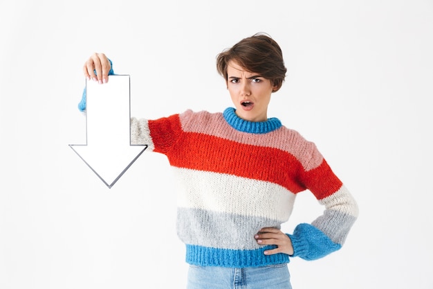 Chica molesta vistiendo suéter de pie aislado en blanco, apuntando hacia abajo con una flecha de papel