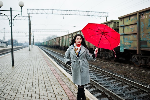 Chica morena en abrigo gris con paraguas rojo en la estación de tren