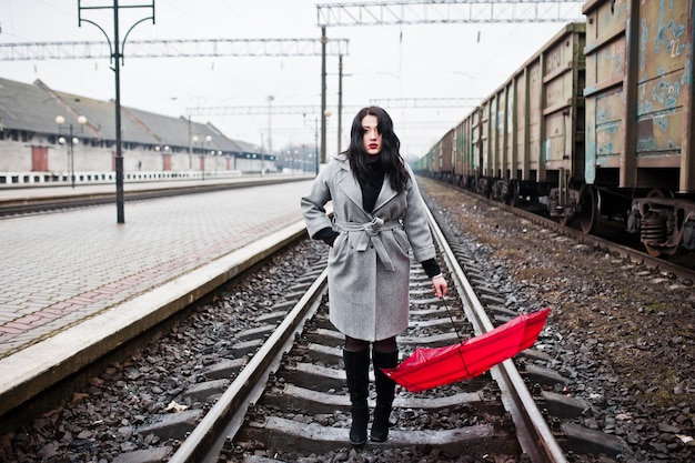 Chica morena en abrigo gris con paraguas rojo en la estación de tren