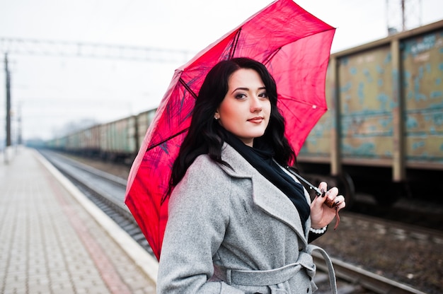 Chica morena con abrigo gris con sombrilla rosa en la estación de tren