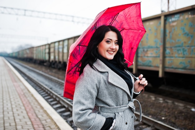 Chica morena con abrigo gris con sombrilla rosa en la estación de tren