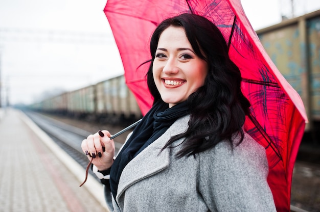 Chica morena con abrigo gris con sombrilla rosa en la estación de tren