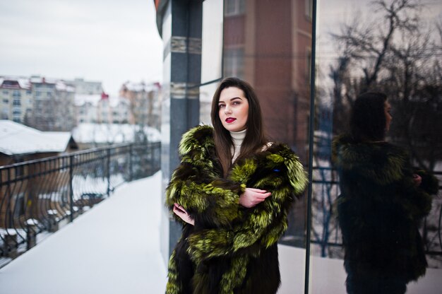 Chica morena en abrigo de piel verde en la calle de la ciudad contra la casa con grandes ventanales en invierno.