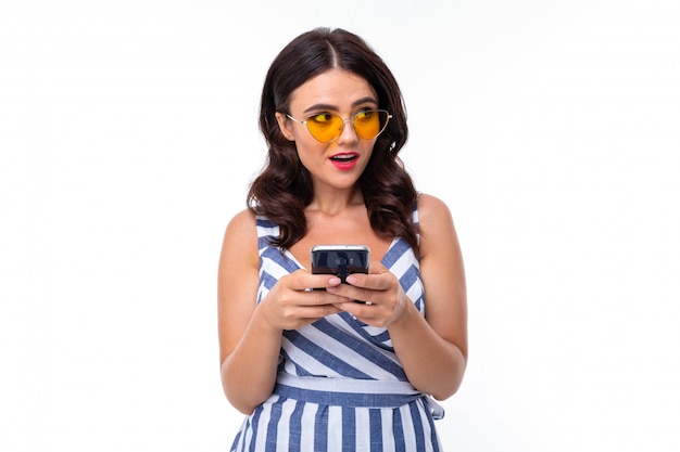Chica morena atractiva en un vestido de rayas y gafas con un teléfono en sus manos sobre una pared blanca