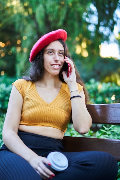 Chica morena de boina roja sentada en el banco del parque hablando por teléfono