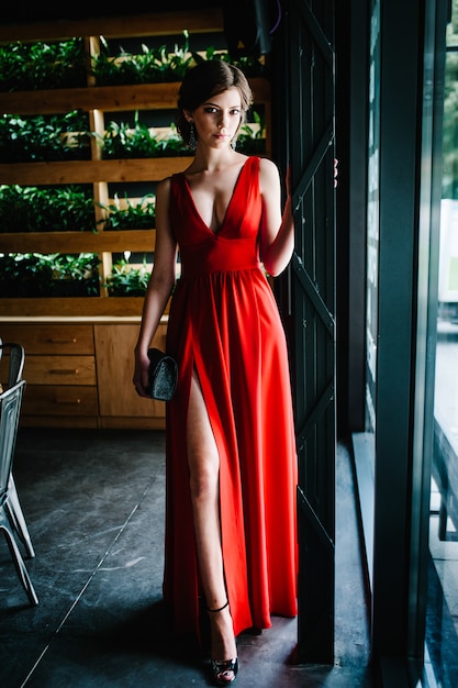 Chica morena con un bolso en la noche vestido rojo para la mujer vestido largo está de pie en el restaurante. | Foto Premium