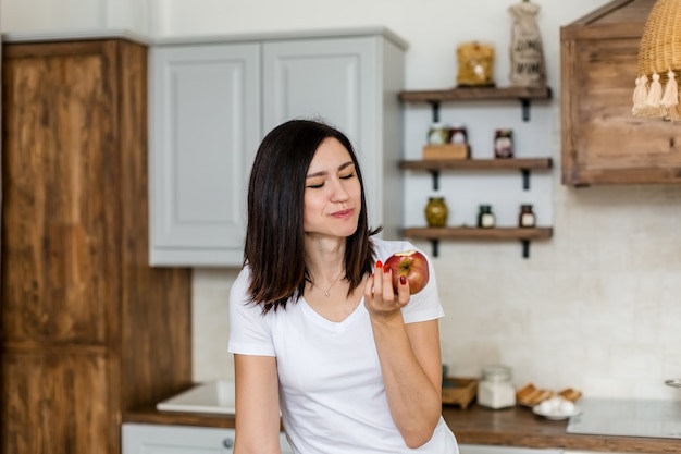 Chica morena con una camiseta blanca en la cocina come una manzana.