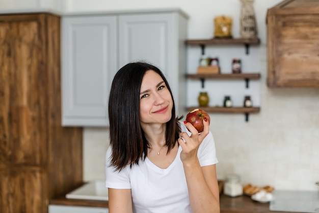 Chica morena con una camiseta blanca en la cocina come una manzana.