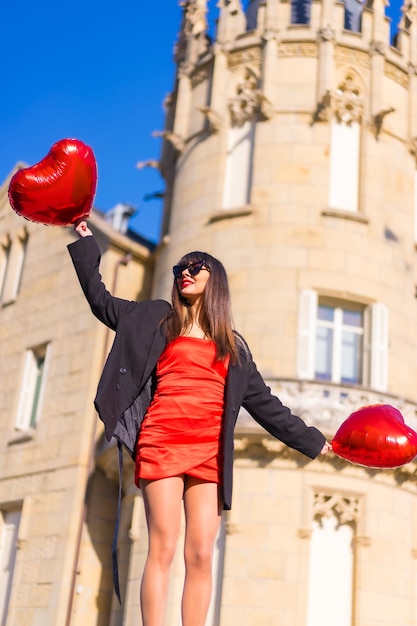 Chica morena caucásica con globos de corazón en el día de San Valentín gafas de sol y vestido rojo Estilo de vida sonriendo y divirtiéndose en un castillo