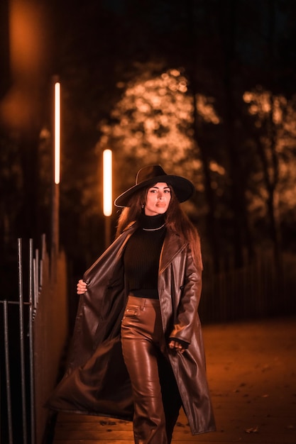 Chica morena caucásica con sombrero caminando por la noche en un parque. Moda de noche de invierno, foto vertical.