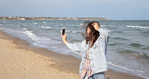 Chica morena con una chaqueta de mezclilla hace una foto en un teléfono con cámara selfie con el fondo del mar. concepto de viajes y nuevas experiencias.