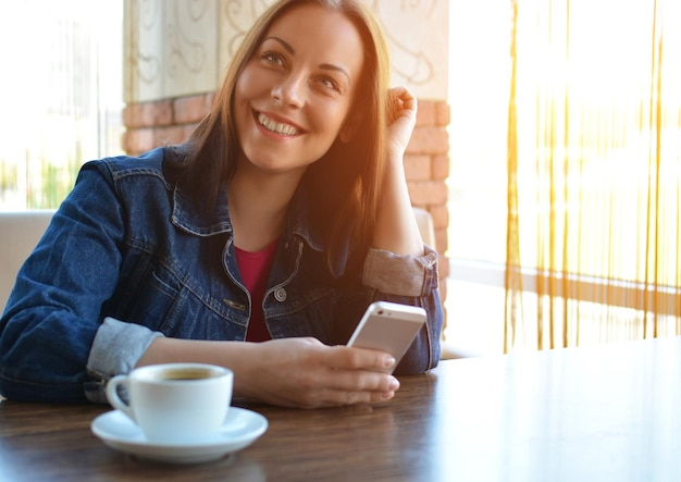 Una chica morena con una chaqueta vaquera está almorzando en un café y usando su teléfono